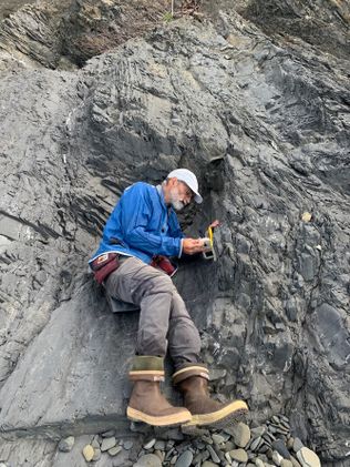 Male wearing a blue long sleeve shirt, white cap, and thick boots perches on a rockface taking sample for use in research